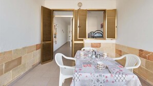 A dining area with a table set for four, white chairs, and a tablecloth with a floral pattern. There are wooden shutters on the walls and a small shelf with items on it.