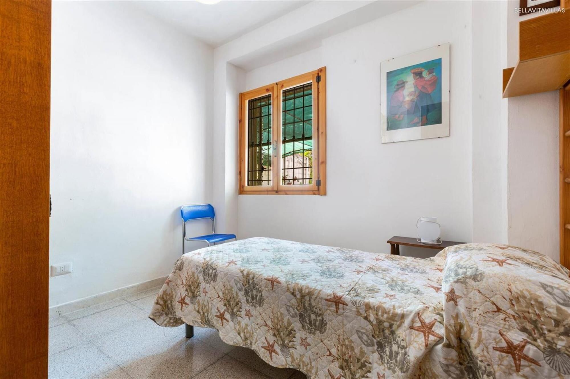 Ein kleiner Raum mit einem Bett, einem Stuhl, einem Fenster mit Gitterstäben und einem Bild an der Wand.