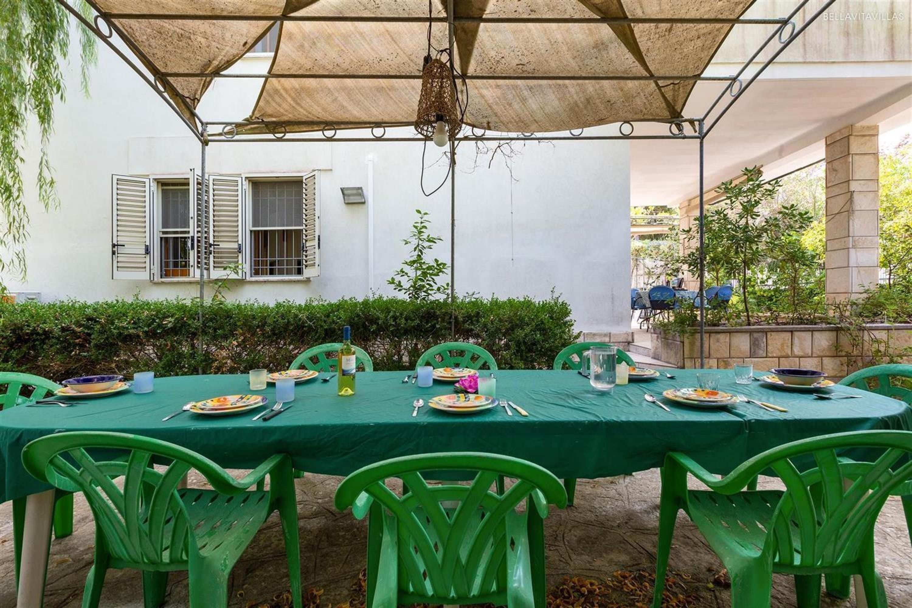 Uma área de refeições ao ar livre com uma mesa posta para seis pessoas, cercada por cadeiras verdes, sob um toldo sombreado.