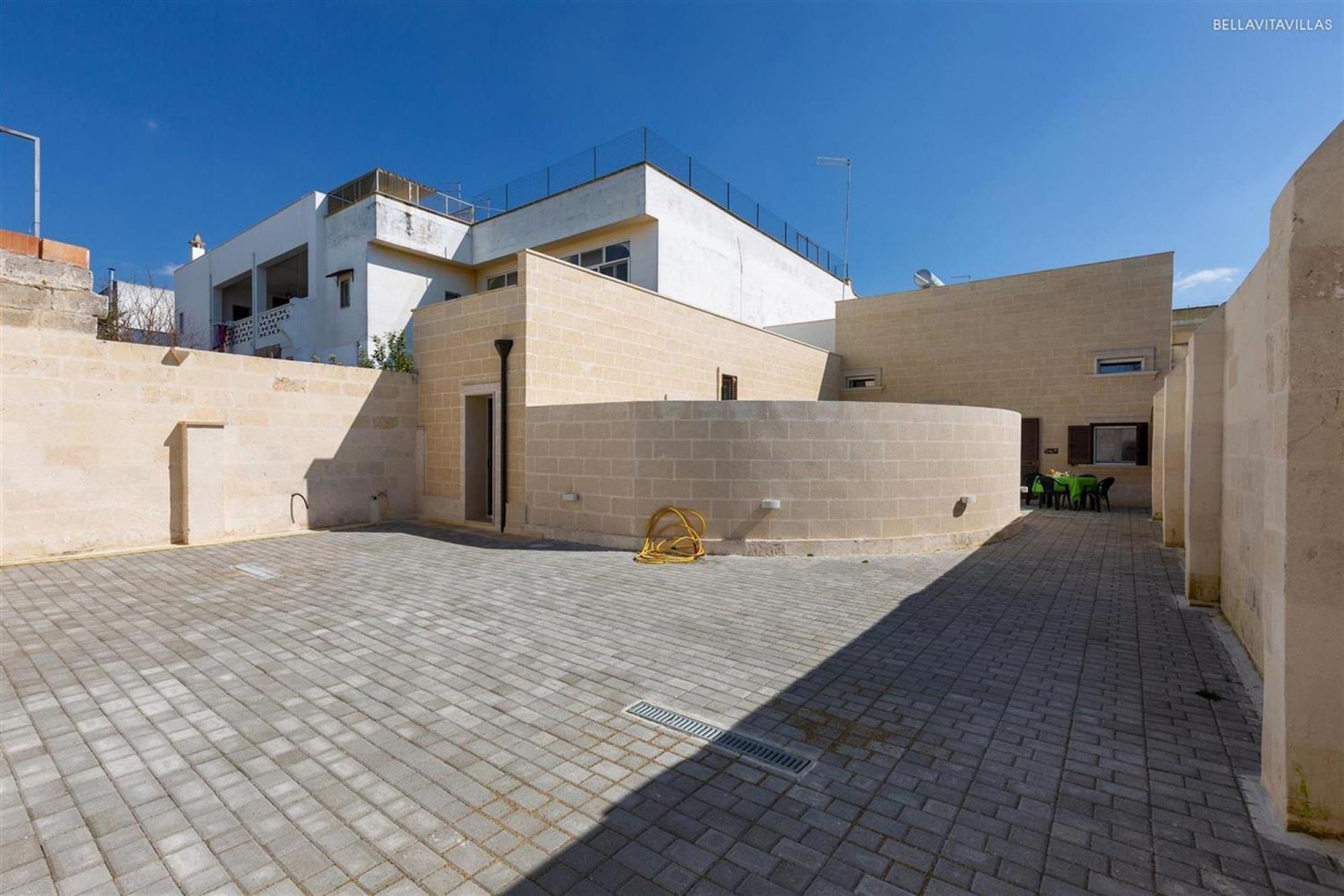 A courtyard with a paved floor, a building with a balcony, and a small table with chairs.