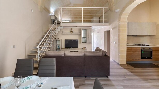 Ein großzügiger Wohnbereich mit hoher Decke, einer Couch, einem Esstisch mit Stühlen, einer Küche mit modernen Geräten und einer Treppe, die zu einer oberen Ebene führt.
