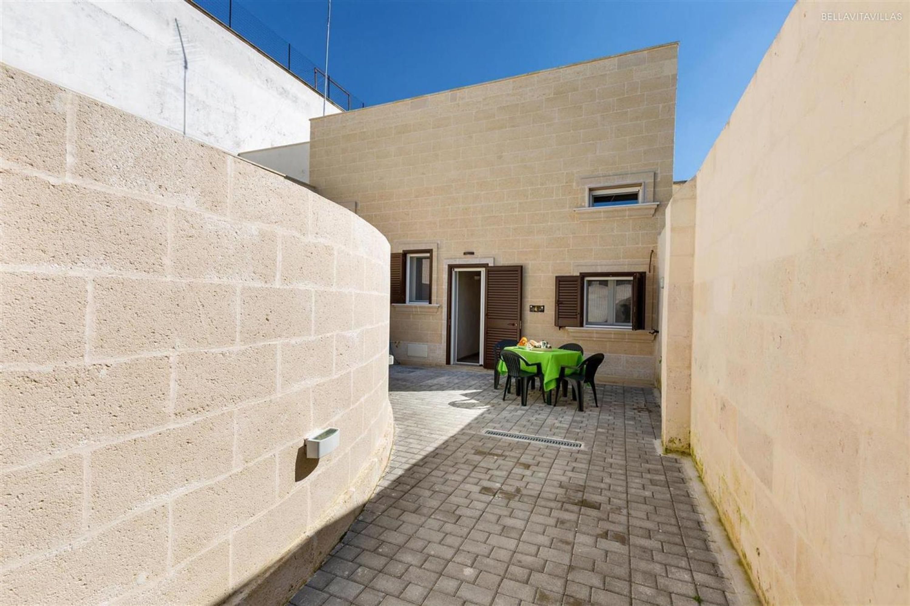 Ein Innenhof mit Backsteinpflaster, einem Tisch mit grüner Tischdecke und einem Gebäude mit beige gefärbten Wänden und Fenstern.