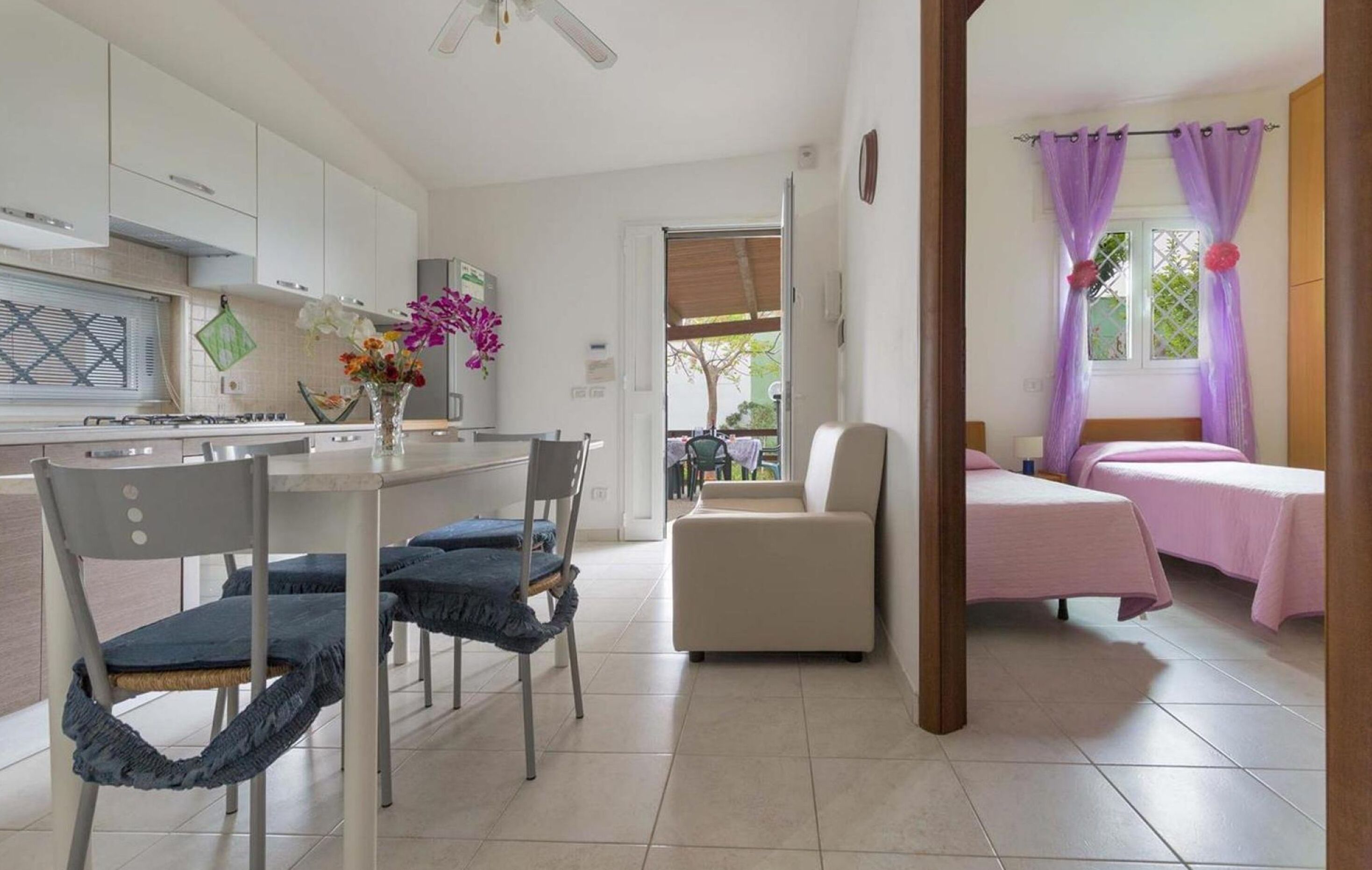 A modern kitchen with white cabinets, a dining table with chairs, a vase with flowers, and a view of a balcony with a chair.