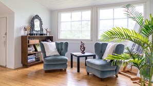 Un salon avec deux fauteuils bleus, une petite table, une bibliothèque et une grande fenêtre.