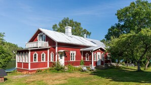 Une maison en bois rouge avec un balcon blanc, entourée d’une cour verdoyante et d’arbres.