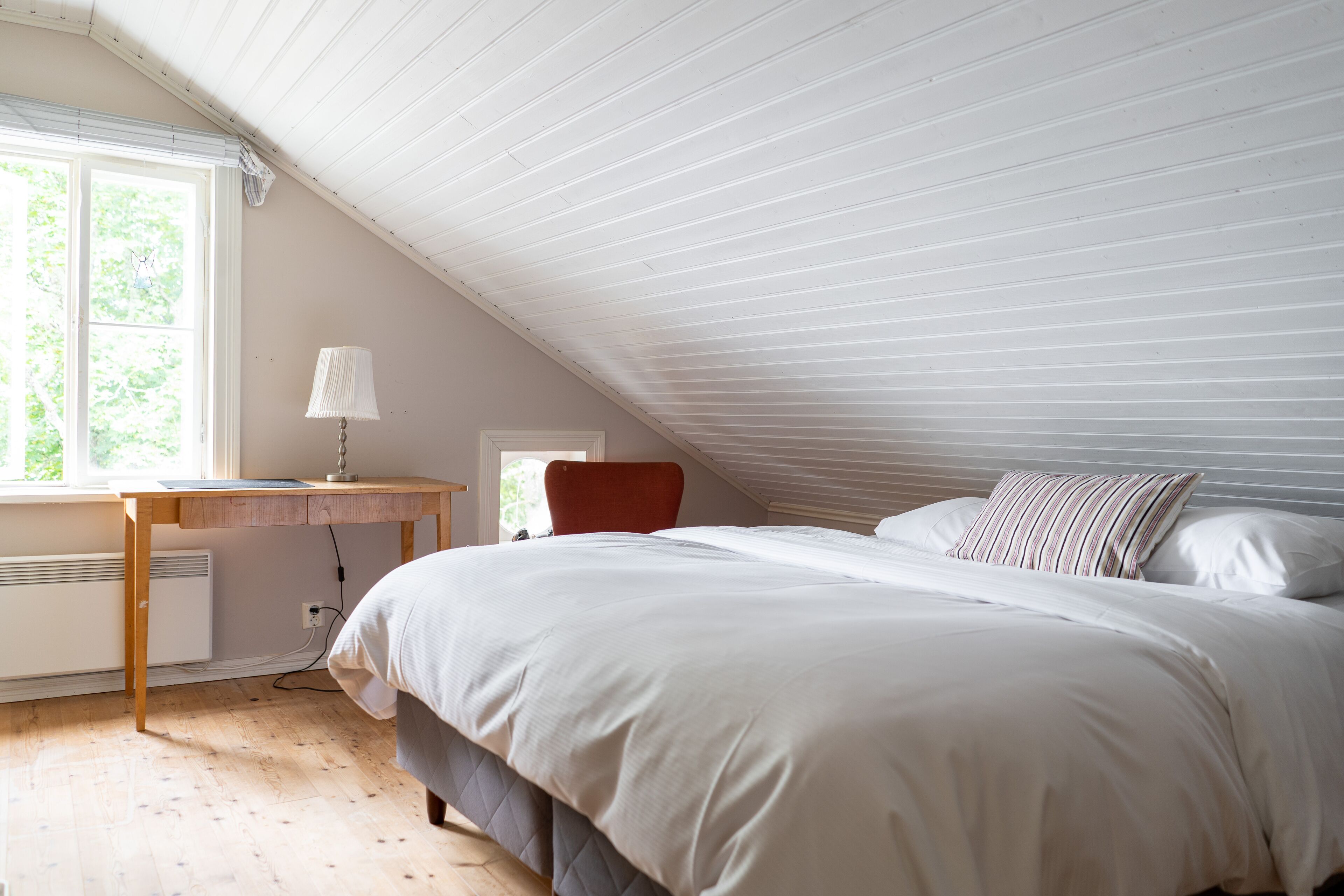 A bedroom with a sloped ceiling, a bed with white bedding, a desk with a lamp, and a window with blinds.