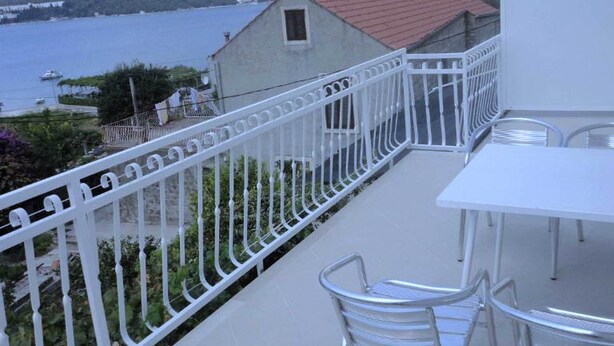 Ein Balkon mit weißer Reling, der auf ein Gewässer und Hügel blickt. Auf dem Balkon stehen ein Tisch und zwei Stühle.