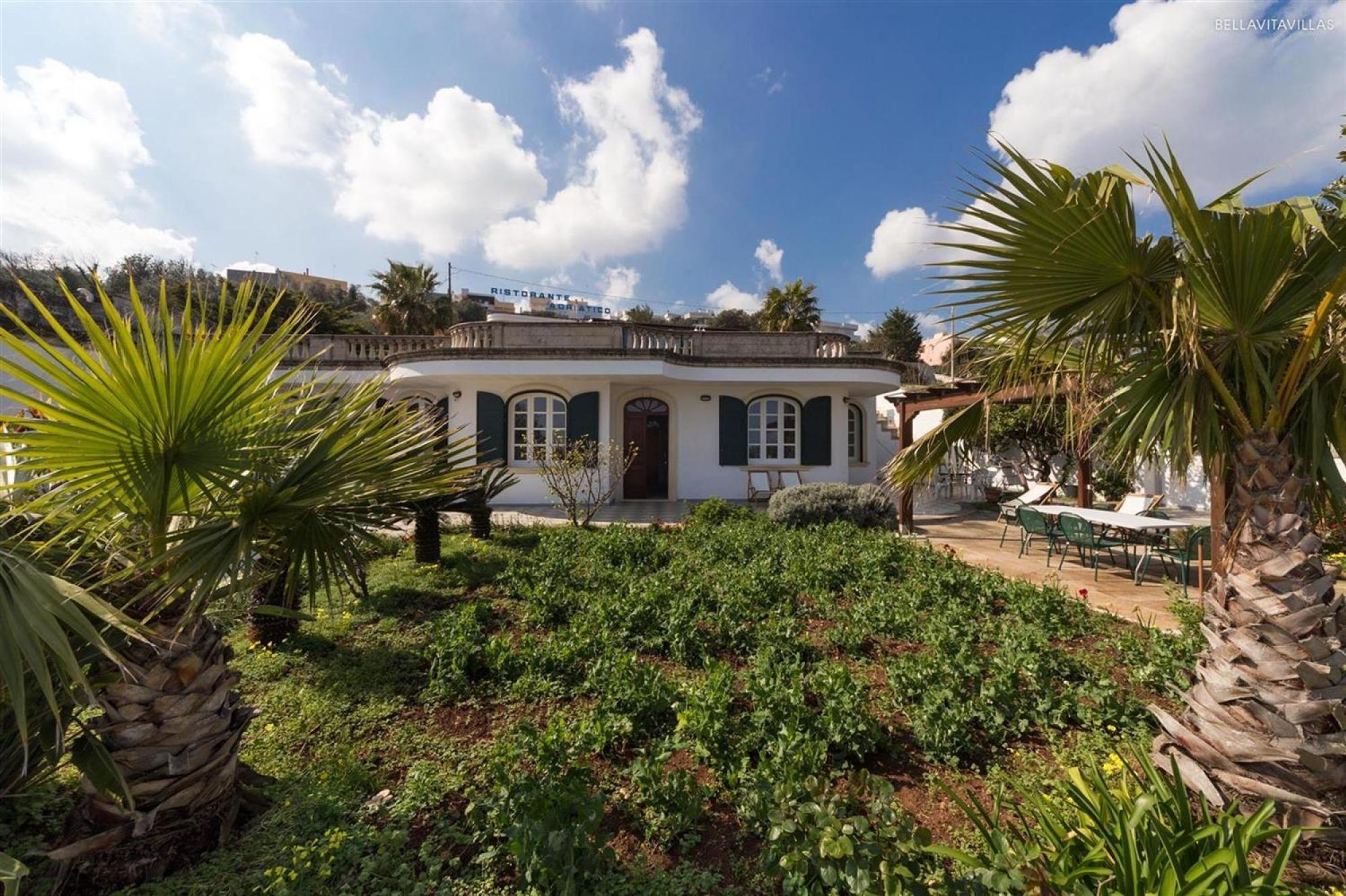 A white building with a green door and windows, surrounded by lush greenery and palm trees.