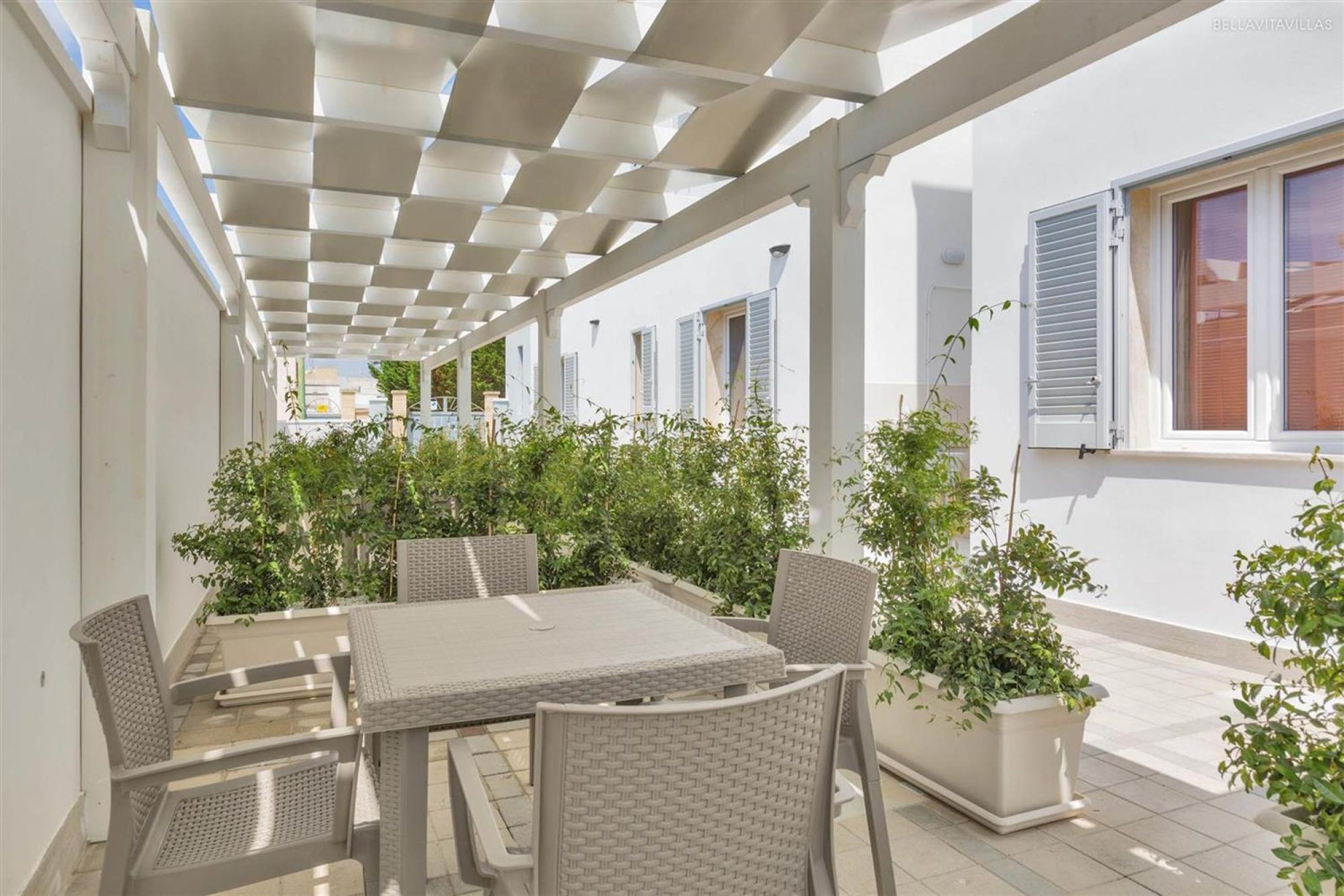 An outdoor patio with a table and chairs, surrounded by potted plants and a pergola structure.