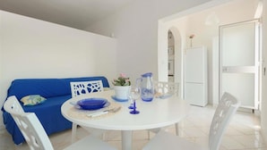 A dining area with a white table, blue chairs, and a blue sofa. There is a white door and a white refrigerator in the background. - 1542 Villetta Playa De Mar (Manduria)