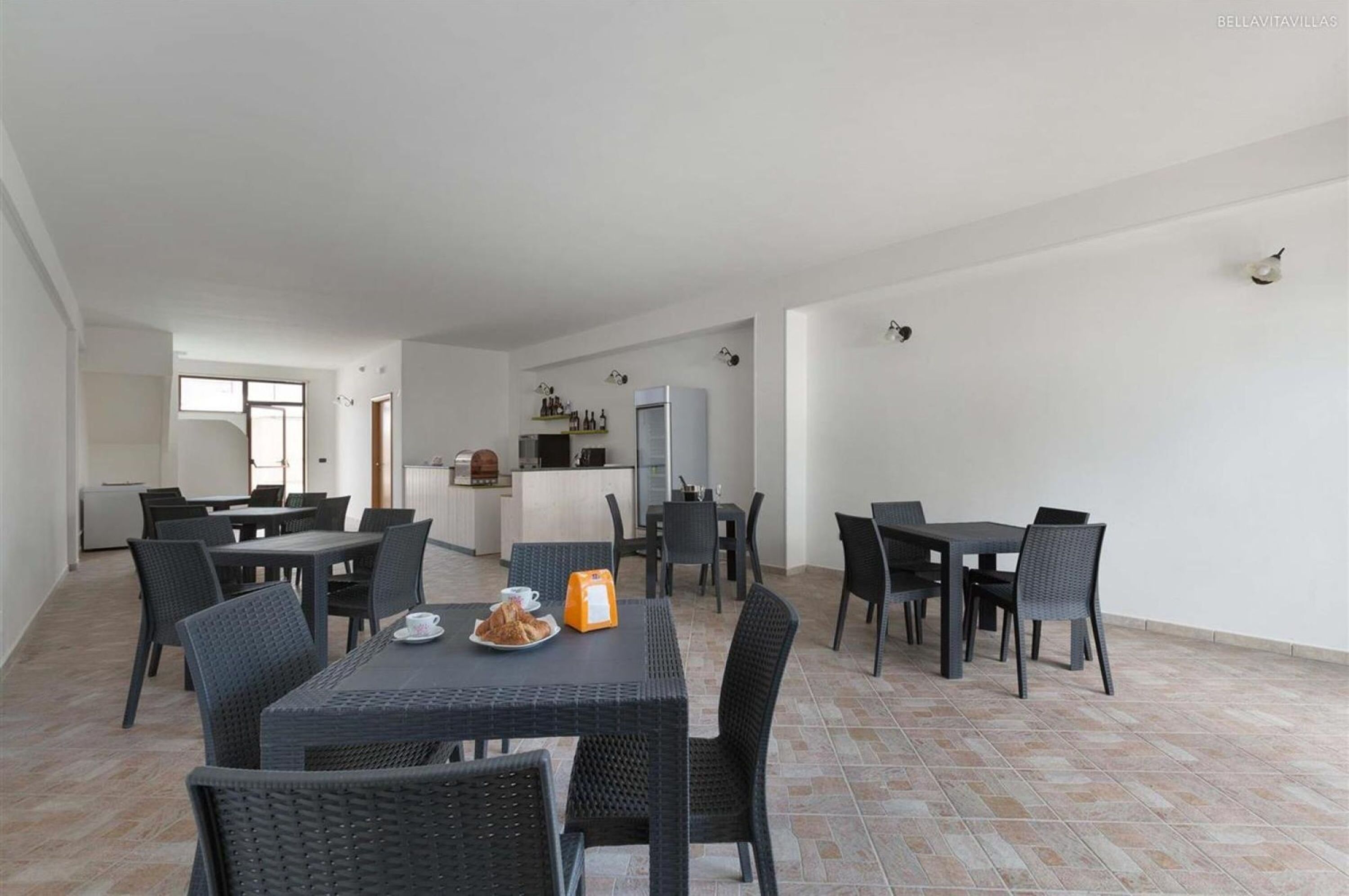 Un grand espace dédié à la salle à manger, avec plusieurs tables et chaises, une cuisine à l’arrière-plan et un sol carrelé.