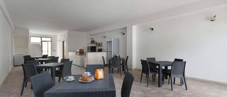 Un grand espace dédié à la salle à manger, avec plusieurs tables et chaises, une cuisine à l’arrière-plan et un sol carrelé.