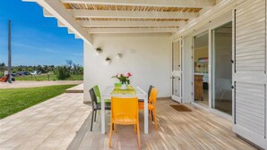 Eine überdachte Terrasse mit Tisch und Stühlen, eine Glastür zu einem Innenbereich und Blick ins Freie.