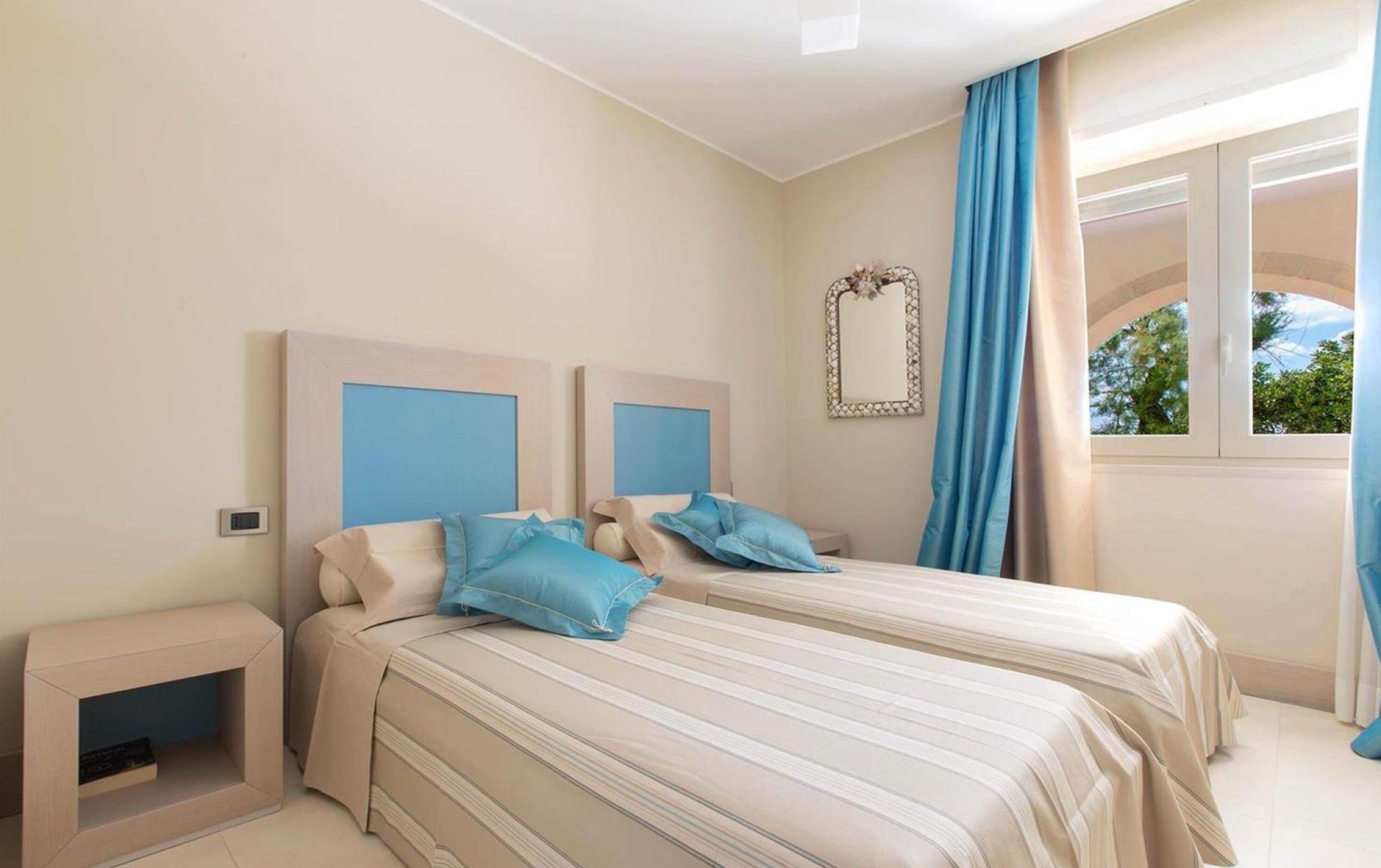 A bedroom with two beds, a bedside table, a mirror, and a window with curtains.