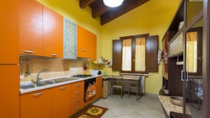 A compact kitchen with orange cabinets, a gas stove, and a sink. There is a dining area with a table and chairs. - 425 Casa in Campagna con Piscina a San Donaci (San Donaci)