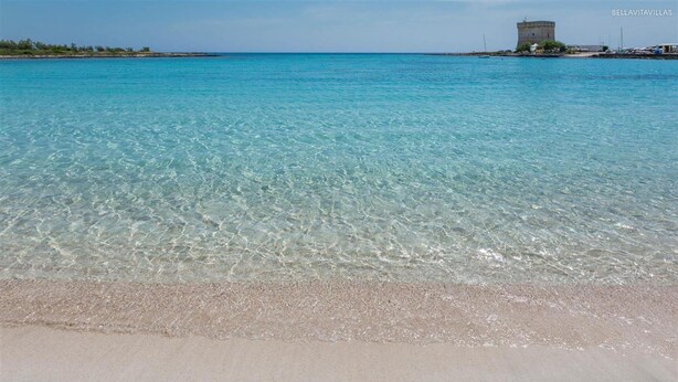 Ein Strand mit klarem türkisblauem Wasser, einem sandigen Ufer und einem Gebäude in der Ferne.