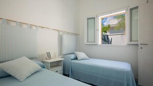 A bedroom with two beds, a window with a view of greenery, a bedside table with a lamp, and a door. - 1687 Villa Blu Cobalto - Appartamento 1 (Manduria)