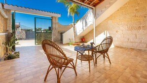 Eine Terrasse im Freien mit Korbstühlen und einem Tisch, einer Steinmauer sowie Blick auf ein Tor und eine Palme.