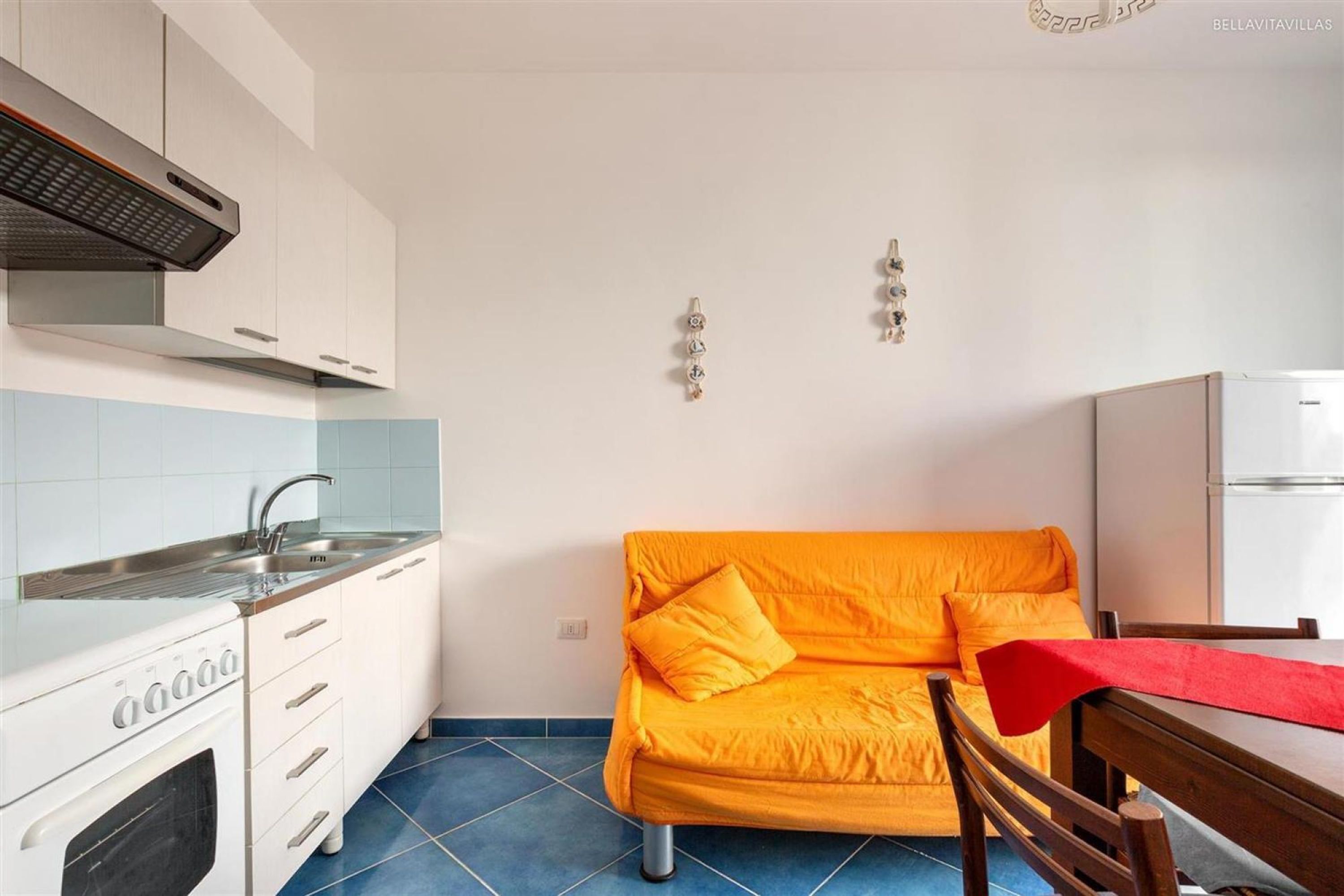A compact kitchen with white cabinets, a built-in oven, and a sink. A bright orange sofa is placed against the wall, and a small dining table with a red tablecloth is visible.