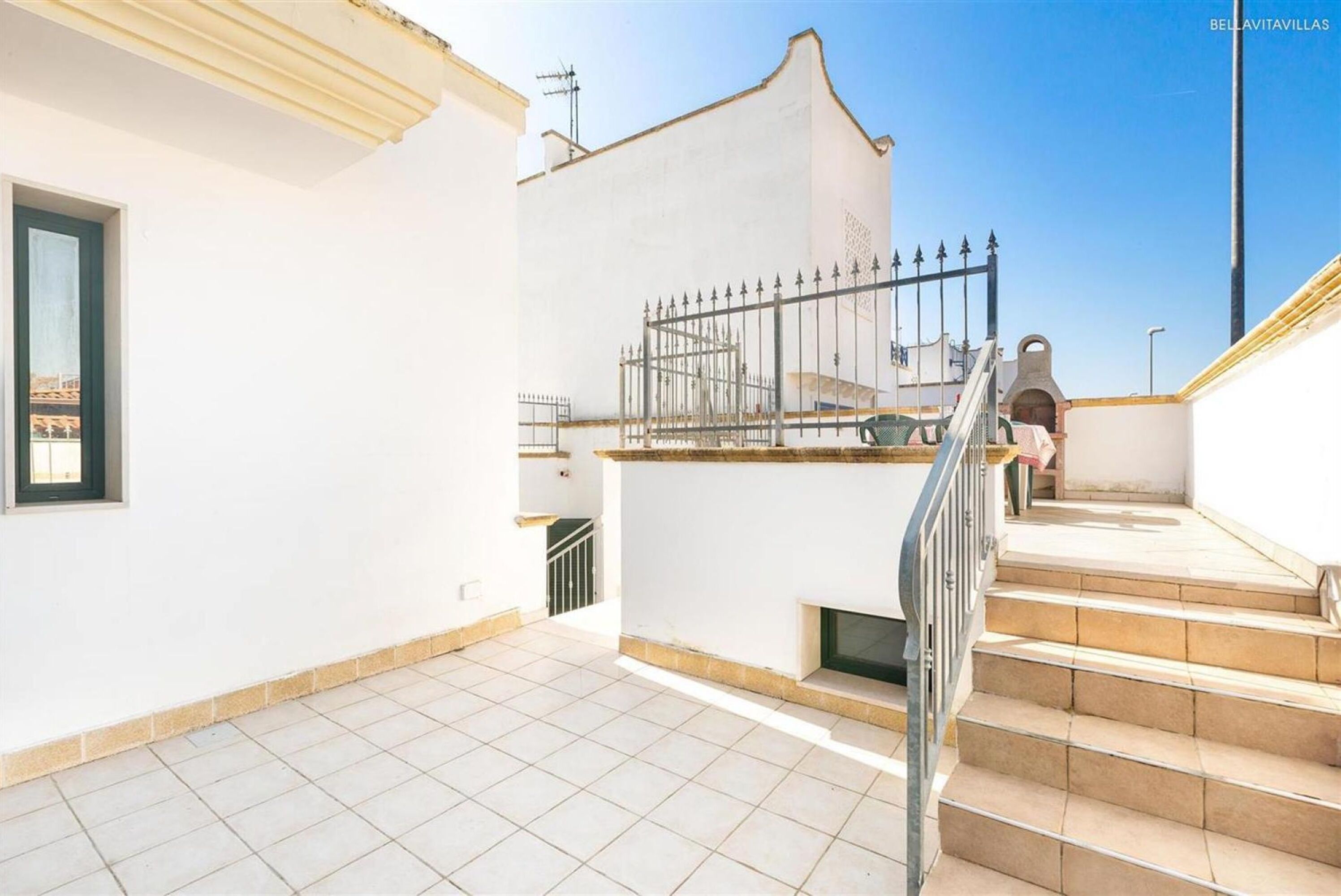 Un bâtiment blanc doté d’un patio carrelé, d’un portail en métal et d’un escalier menant à un étage supérieur.