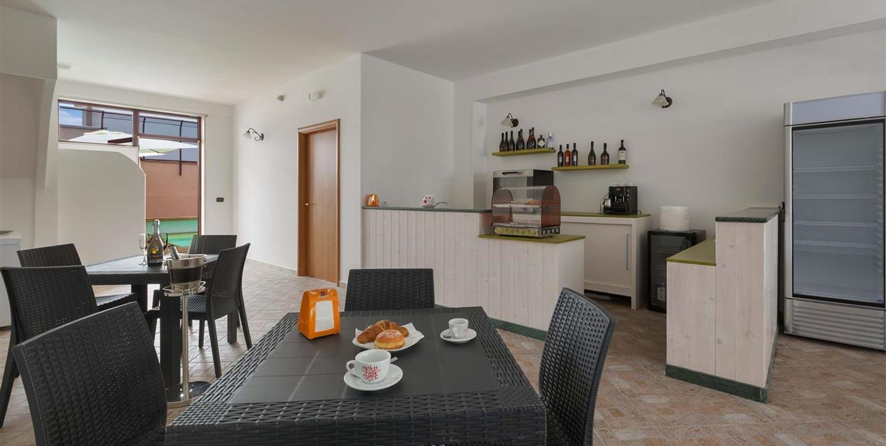 Eine moderne Küche mit Kaffeemaschine, Kühlschrank und einer Ablage mit Flaschen. Es gibt eine Essecke mit Tisch und Stühlen sowie eine Tür zu einem anderen Raum.