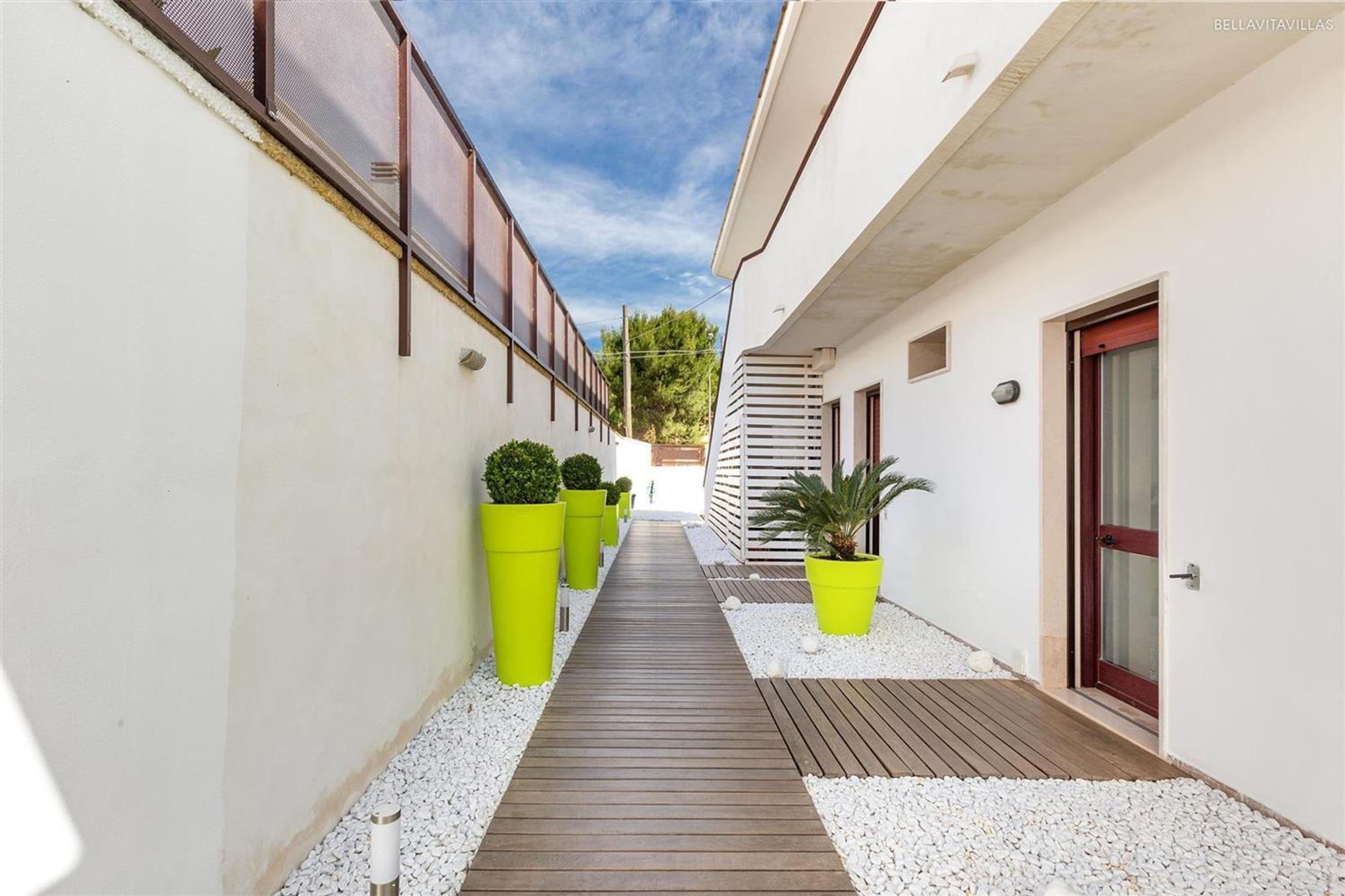 A narrow pathway with wooden decking, flanked by white walls and green planters.