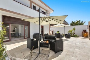 Eine moderne Terrassenanlage im Freien mit Korbmöbeln, einem großen Sonnenschirm und einem Essbereich, der durch eine Glastür sichtbar ist.