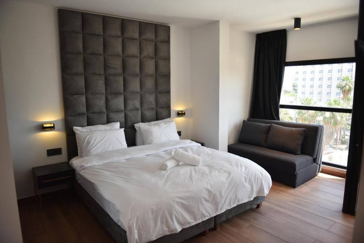 A modern hotel room with a large bed, a dark sofa, and a large window offering a view of buildings and greenery.