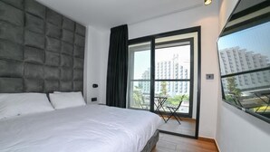 Een moderne slaapkamer met een groot bed, een grijs hoofdeinde, een balkon met een tafel en uitzicht op de stad.