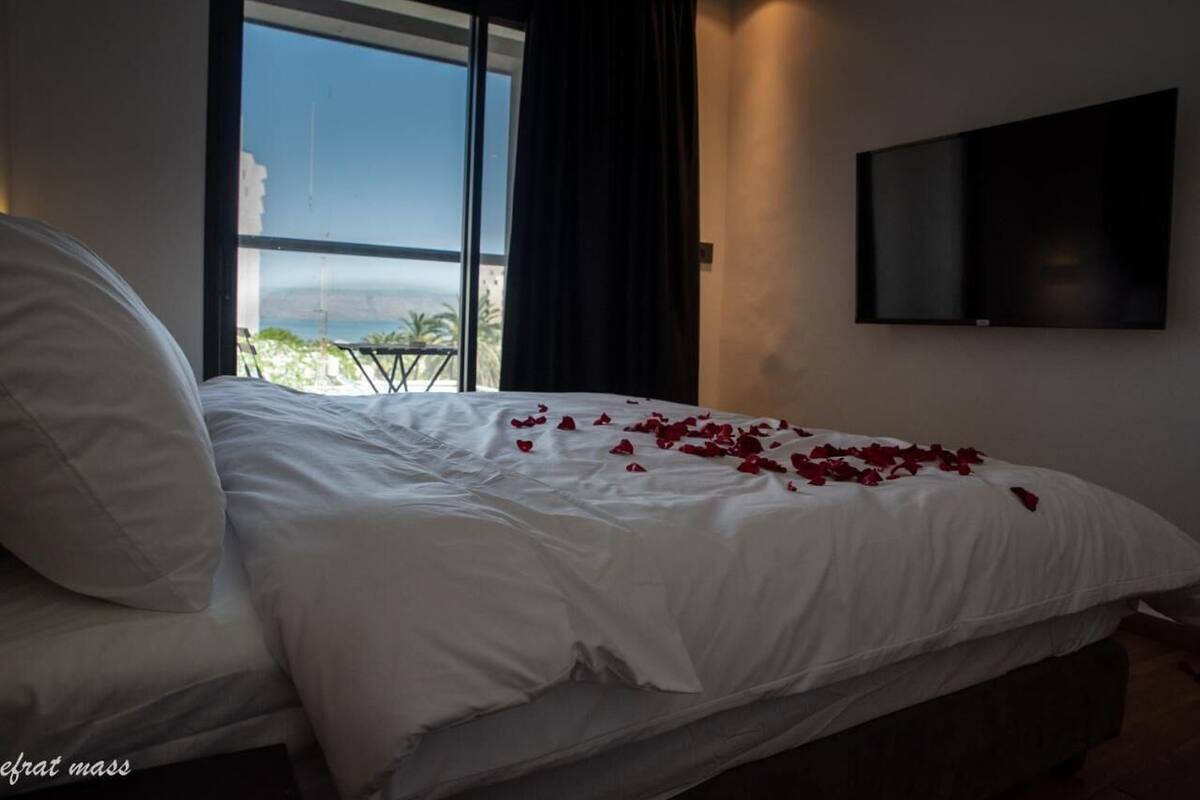 A bed with white linens and rose petals scattered on it, a large window with a view of the sea, and a flat-screen TV mounted on the wall.