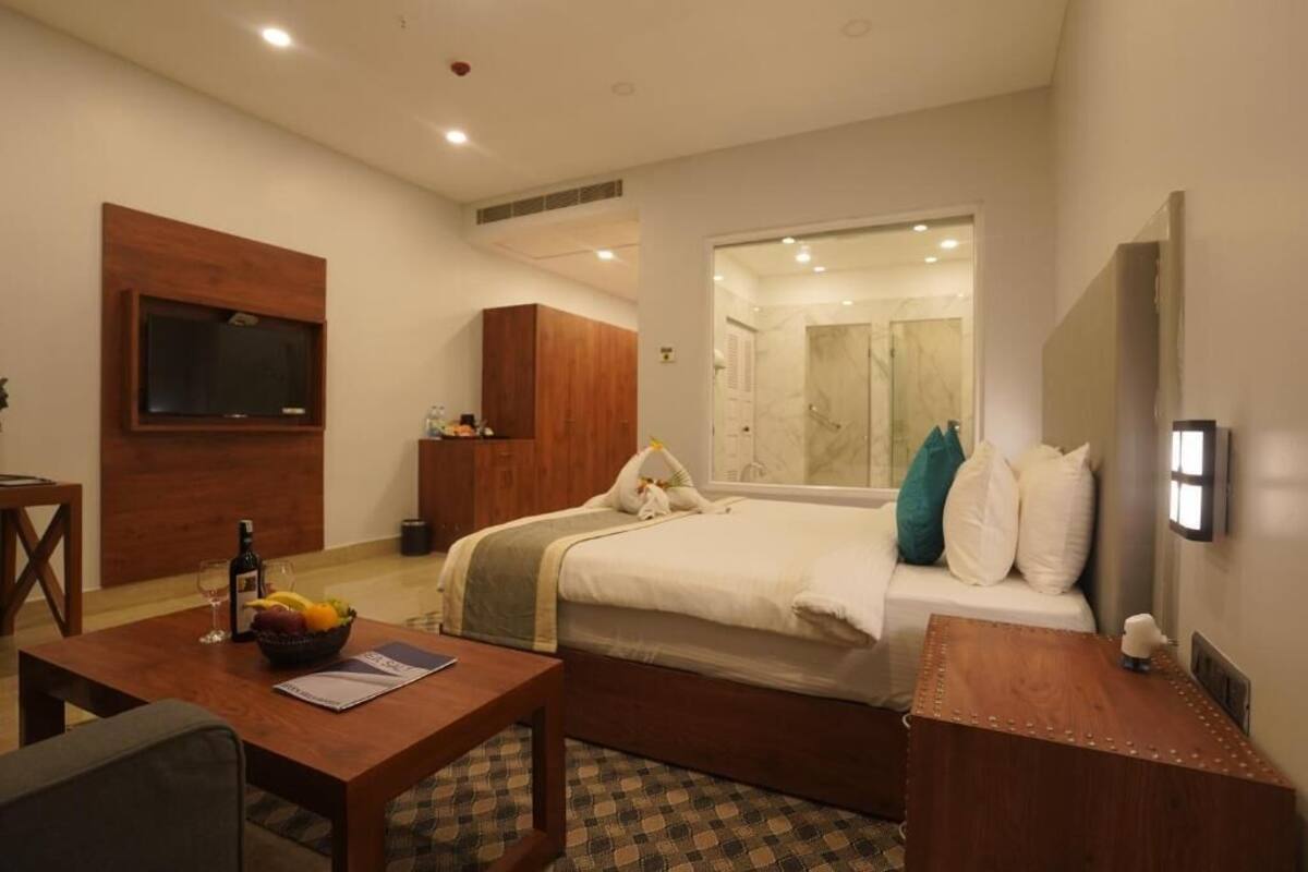 Habitación de hotel con cama, escritorio con silla, televisión y baño visible a través de una puerta de cristal.