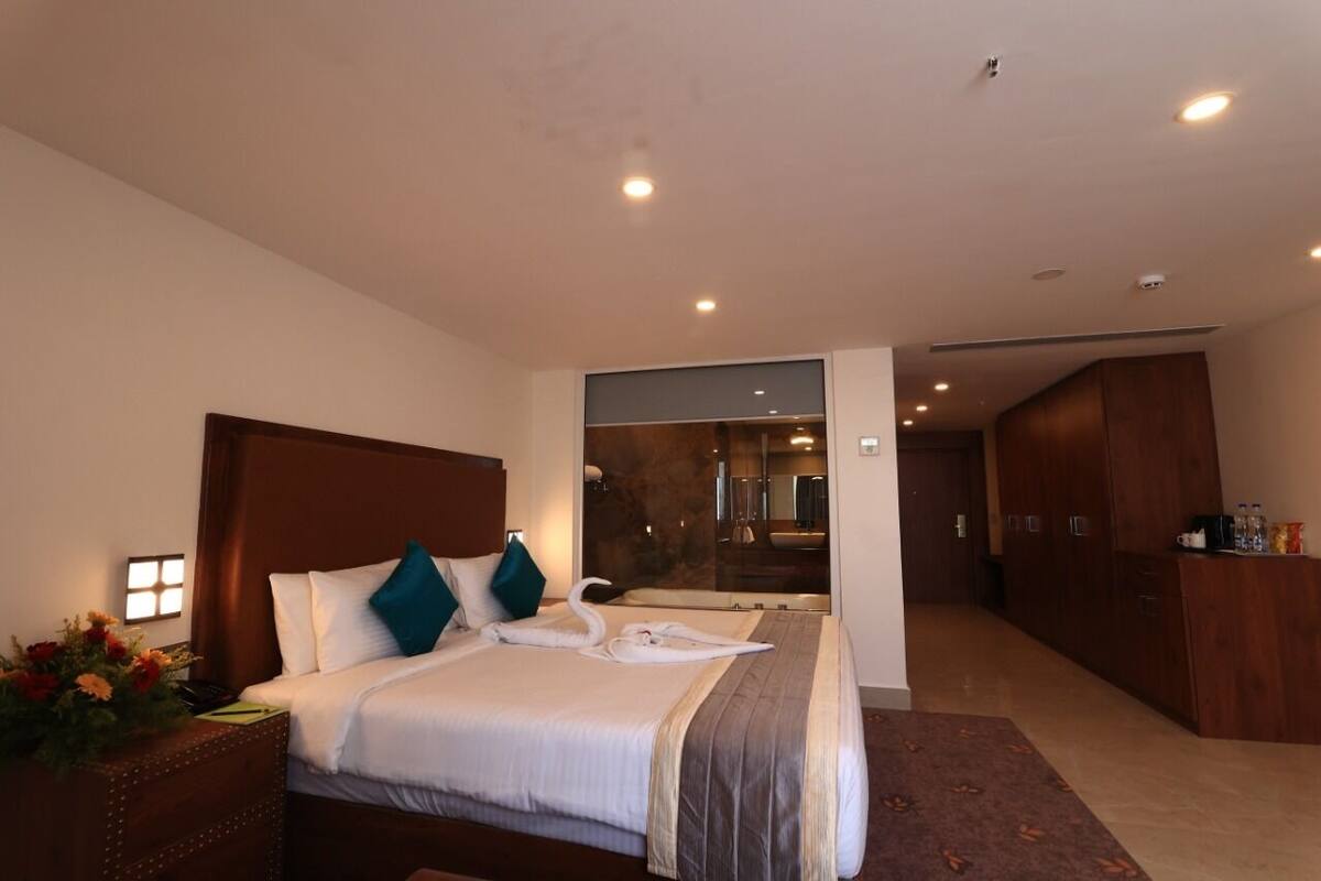 Una habitación de hotel con una cama grande, un armario de madera, un baño con puerta de vidrio y un pequeño escritorio con un florero.