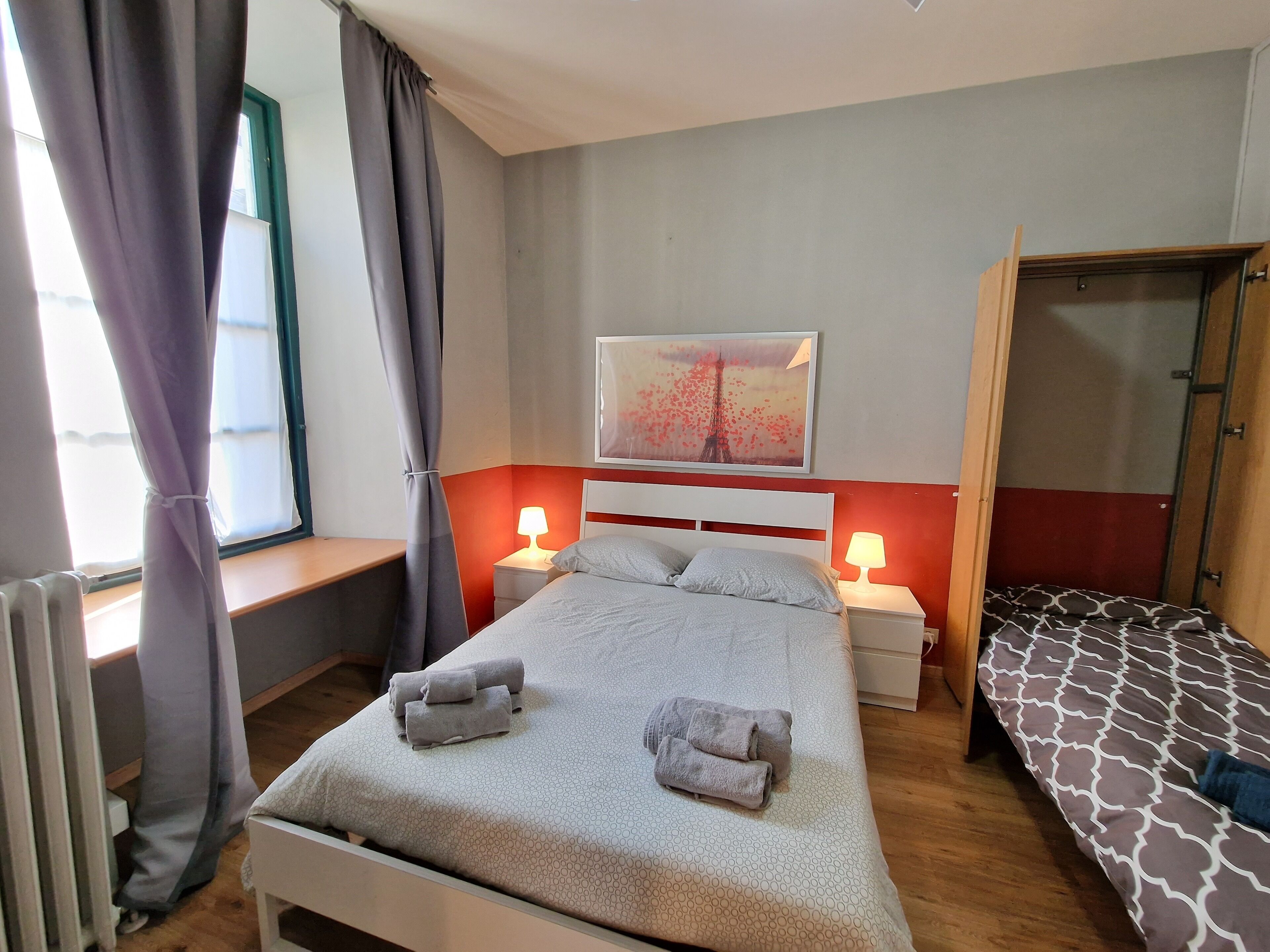 Une chambre d’hôtel avec un lit, deux tables de chevet avec des lampes, une fenêtre avec des rideaux, un tableau représentant la Tour Eiffel et une armoire en bois.