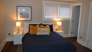 A bedroom with a dark blue bedspread, two bedside lamps, a framed picture above the bed, and a window with blinds. - Charming renovated historic home in ideal location (Hamilton)