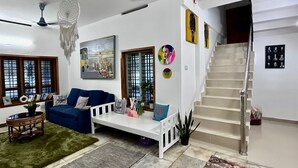 A living room with a blue sofa, a white bench, a patterned rug, a ceiling fan, and a staircase. - Home in Varkala (Varkala)