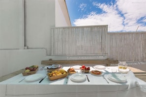 A table set with plates, bowls of fruit, and a glass of water on a terrace with a white wall and a clear sky in the background.