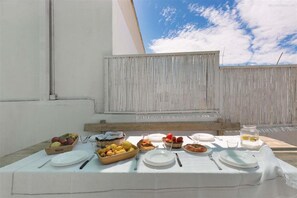 A table set with plates, cutlery, and bowls of fruit and food on a terrace with a white wall and a clear sky in the background.