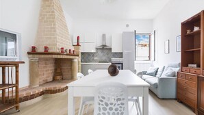 A modern kitchen with a brick fireplace, white cabinetry, and a dining area with a white table and chairs. - 857 Appartamento Sirena (Porto Cesareo)