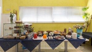 Mesa com buffet de café da manhã, diversos alimentos e bebidas, máquina de café e vitrine com doces.