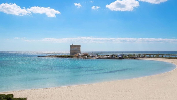A beach with clear turquoise water, a sandy shore, and a small building structure in the distance. - 2091 Villino Tropical (Porto Cesareo)