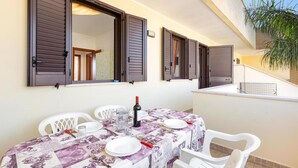 Un coin repas avec une table dressée pour quatre personnes, recouverte d’une nappe à motifs, et quatre chaises blanches. On peut voir trois fenêtres avec des volets marron, et une plante en pot est visible à l’arrière-plan.