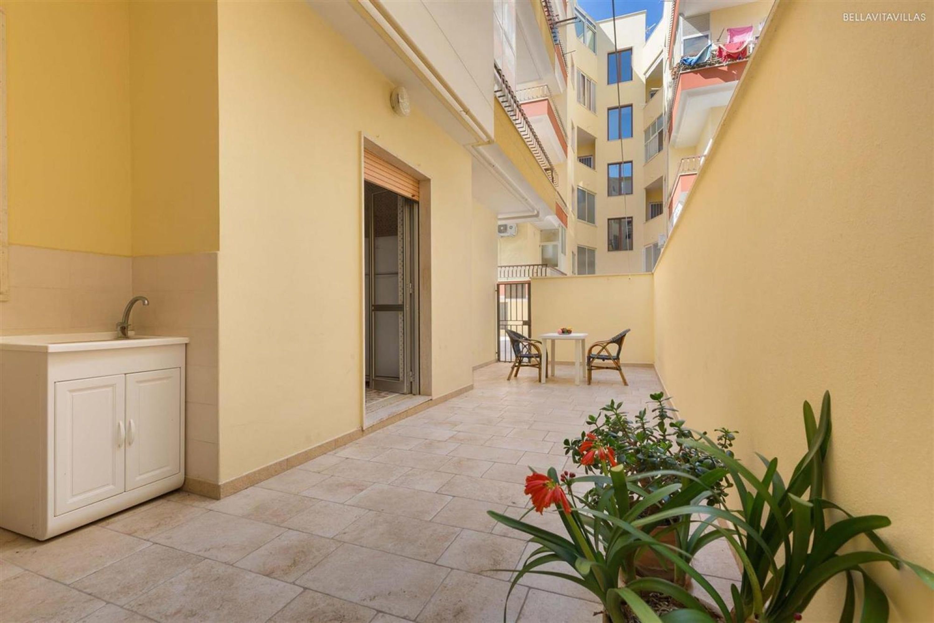 A narrow corridor with a sink and cabinets, a small table with chairs, and potted plants.