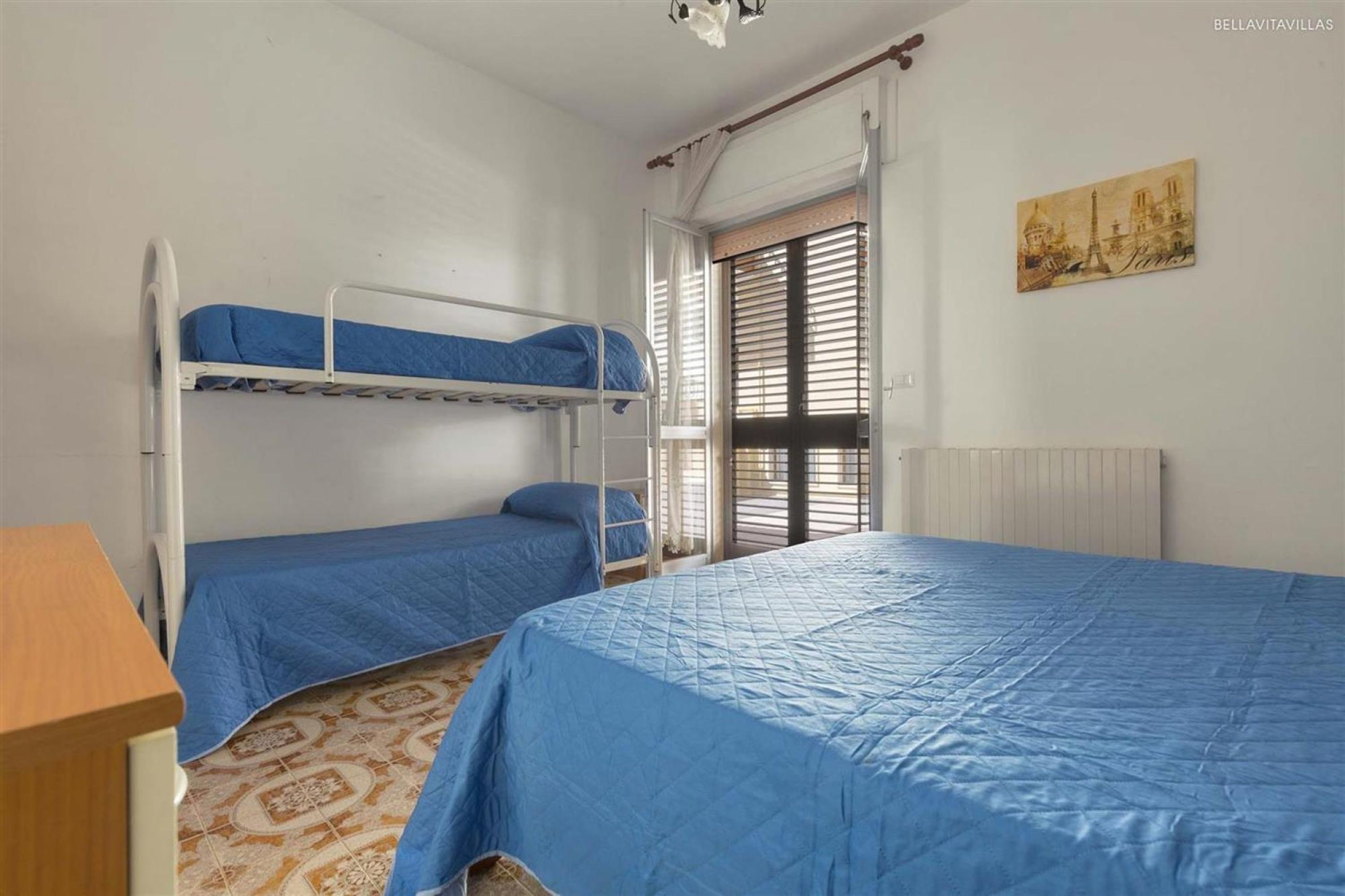 Une pièce avec des lits superposés, un radiateur, une fenêtre avec des stores et un tableau accroché au mur.