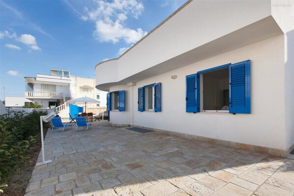 Un bâtiment blanc avec des volets bleus, une terrasse pavée avec des chaises et un ciel bleu clair.