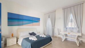 A modern hotel room with a bed, a desk, and a chair. There is a painting of a beach on the wall.