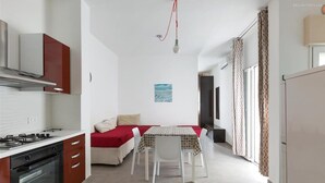 A compact kitchen with a stove, oven, and cabinets, a dining area with a table and chairs, and a bed with a red blanket. - 1299 Versomare - Monolocale Lu Mare (Porto Cesareo)