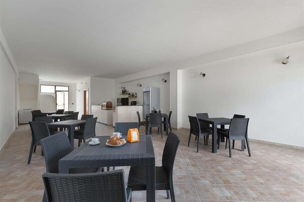 Un amplio comedor con varias mesas y sillas, una cocina al fondo y piso de baldosas.