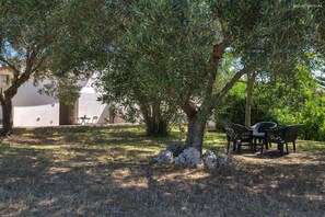 Ein Sitzbereich im Freien mit Tisch und Stühlen unter einem Baum, umgeben von einer grünen Rasenfläche und einem Gebäude mit weißer Fassade.