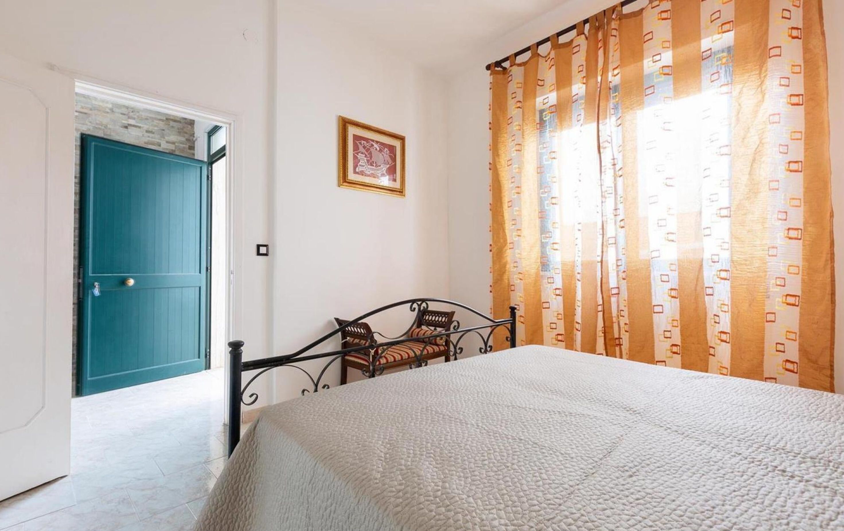 A bedroom with a bed, a window with curtains, a door, and a framed picture on the wall.