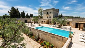 Zona de piscina con una piscina azul cristalina, rodeada por un muro de piedra y diversas plantas. Hay sillas de descanso y un edificio con una estructura tipo torre al fondo.